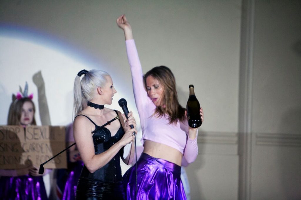 A blonde woman holding the mic and the other in raising right her and and her left hand holding a wine bottle.