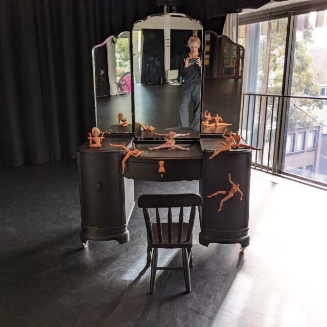 Seven barbie dolls scattered on a black dressing table with three mirrors with a child-size black chair in front. In the reflection of the mirror is Bonnie taking a photo.